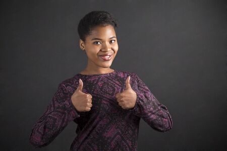 South African or African American black woman teacher or student with a thumbs up hand signal standing against a chalk blackboard background insideの写真素材