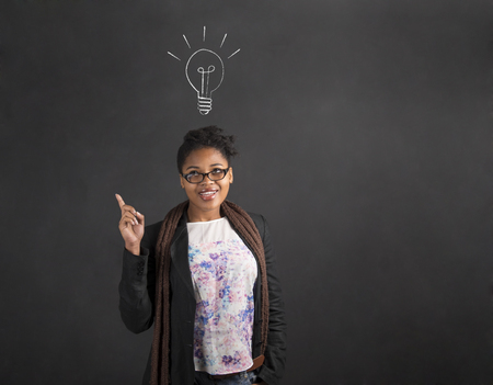South African or African American black woman teacher or student with a good idea or answer lighbulb standing against a chalk blackboard background insideの写真素材