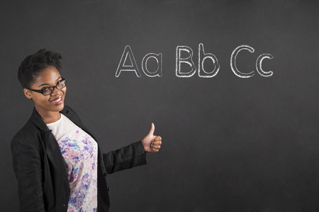South African or African American black woman teacher or student with a thumbs up hand signal showing the alphabet standing against a chalk blackboard background insideの写真素材