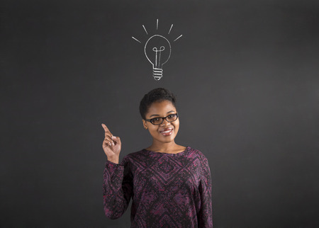 South African or African American black woman teacher or student with a good idea or answer lighbulb standing against a chalk blackboard background inside