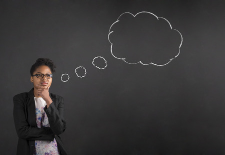 South African or African American black woman teacher or student with her hand on her chin whilst thinking thought cloud or bubble standing against a chalk blackboard background inside