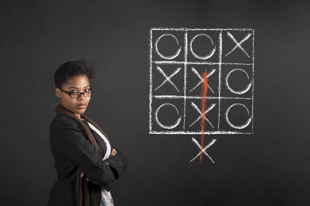 South African or African American woman teacher or student with arms folded tic tac toe diagram on chalk black board backgroundの写真素材
