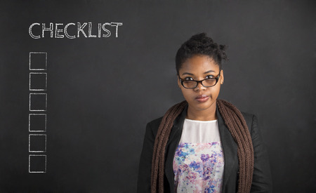 South African or African American black woman teacher or student with her arms behind her back with a checklist on chalk black board background insideの写真素材