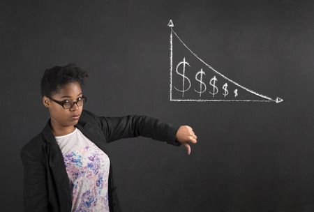 South African or African American black woman teacher or student with thumbs down hand signal to decrease in money on a chalk black board background insideの写真素材