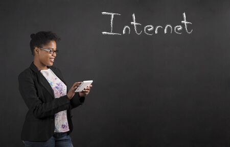 South African or African American black woman teacher or student holding a tablet standing against a chalk blackboard background with Internet word insideの写真素材