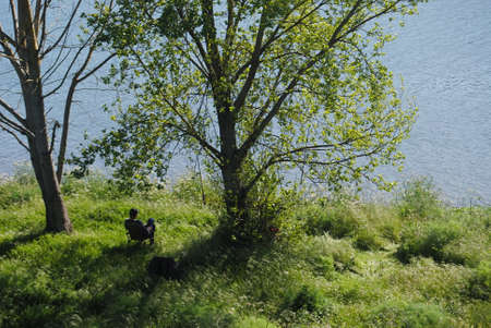 Relaxation at the riverside - man sitting on a chair at the shade of the trees fishing in the riverの写真素材