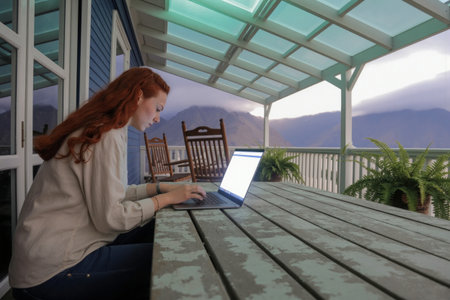 The wonderful free lifestyle of a digital nomad. Young woman working on laptop in cozy environment with mountain view - symbol of work-life balance.の素材