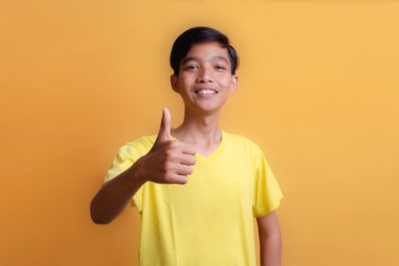 Portrait of Asian young man wearing yellow t-shirt. Thumbs up. show both thumbs. Looking at the camera. Half body portrait on yellow background.の写真素材