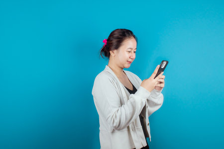 A young Asian woman is happy and smiling while using a smartphone on a blue background. Portrait of a happy Asian woman smiling and holding her smart phone.の写真素材