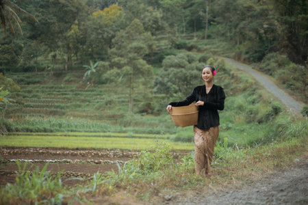 Young woman from an Indonesian village carrying a basket or basket made of woven bamboo. Asian village woman standing and walking in the village rice fields.の写真素材