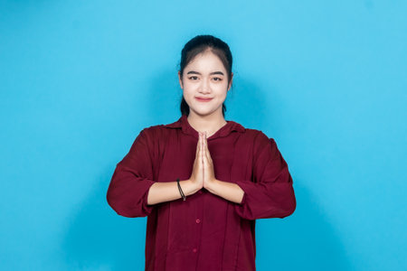 Young woman in maroon posing gracefully, offering a traditional greeting with hands pressed...