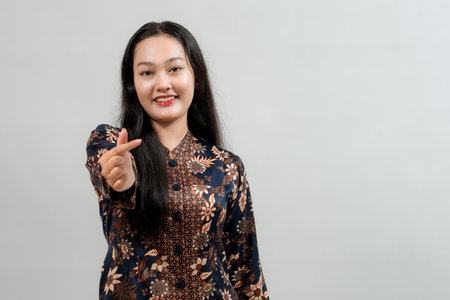 Young Asian woman wearing traditional batik dress, smiling and making a finger heart gesture, isolated on a neutral background.の写真素材
