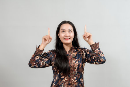 A young woman in beautiful Indonesian batik attire, pointing upwards with a look of wonder and excitement, ideal for showcasing announcements, promotions, or anything overheadの写真素材