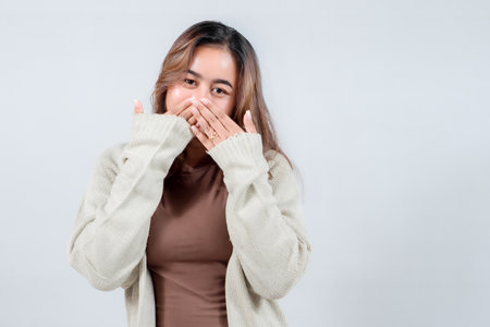A young woman in a cozy sweater covers her mouth with her hands, expressing secrecy, surprise, or...