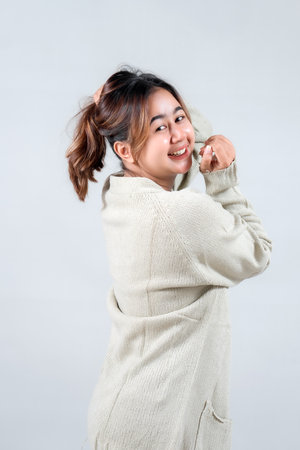 A young woman in a cozy sweater smiles playfully while adjusting her ponytail, radiating a cheerful and casual vibe against a neutral backgroundの写真素材