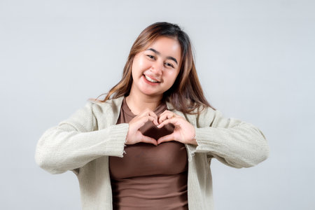 A smiling young woman in a cozy sweater makes a heart shape with her hands, expressing love,...
