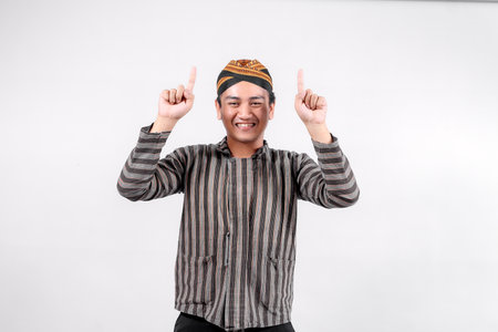 A young man wearing traditional lurik attire smiles brightly while pointing upwards with both hands, showcasing cultural pride and enthusiasm against a neutral background.の写真素材