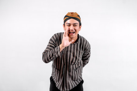 A young man wearing traditional lurik attire smiles widely while gesturing as if calling out, showcasing cultural pride and enthusiasm against a neutral background.の写真素材