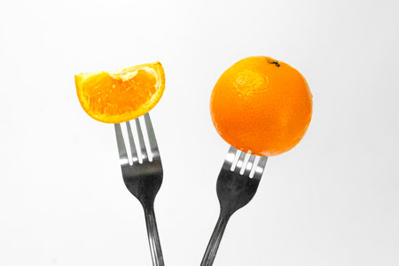 A vibrant orange and a slice of orange are elegantly displayed on forks against a clean white background, showcasing their fresh and juicy appeal.の写真素材