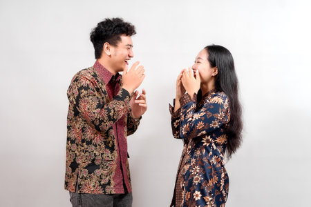 A young couple, dressed in vibrant Indonesian batik shirts, laugh together against a simple gray background. Their joyful expressions and stylish attire showcase Indonesian cultural fashion.の写真素材