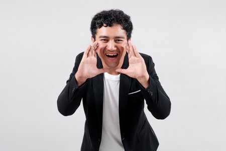 Asian man in black blazer and white t-shirt smiling while shouting with hand near mouth. Isolated on...