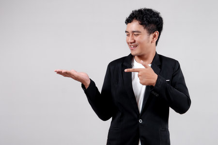 Smiling Asian man in black suit pointing to empty space with open palm, presenting or promoting product. Isolated on white backgroundの写真素材