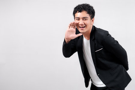 Asian man in black blazer and white t-shirt smiling while shouting with hand near mouth. Isolated on white background. Concept of announcement, calling, or communication.の写真素材