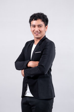 A confident Asian man in a black suit and white shirt stands with arms crossed, smiling slightly. He poses against a plain white background, exuding professionalism and charisma.の写真素材