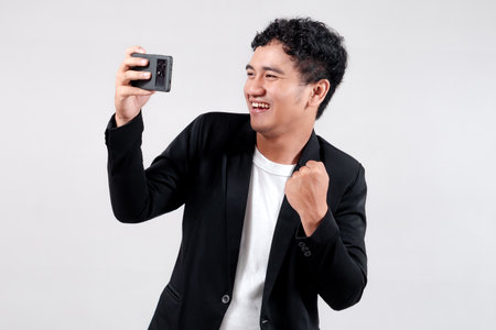 Asian man in a black blazer and white shirt celebrating victory while looking at his smartphone. He is smiling with excitement, raising his fist joyfully in a success gesture.の写真素材