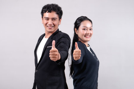 Asian man and woman standing back-to-back, smiling confidently, showing thumbs up. They wear business attire, symbolizing success, teamwork, motivation, and a positive work environment.の写真素材