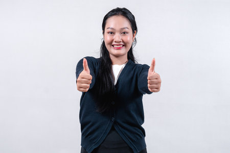 A smiling Asian woman in a black cardigan, with long dark hair, giving a thumbs-up gesture with her hand, against a neutral background.の写真素材