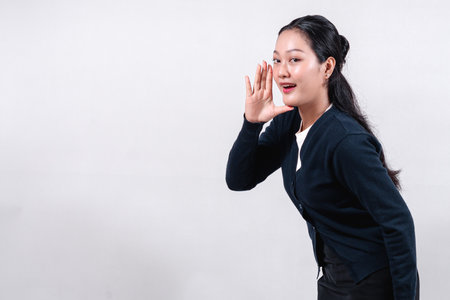 An Asian woman in a black cardigan, with long dark hair, cupping her hand around her mouth as if to whisper, with a playful expression, against a neutral background.の写真素材