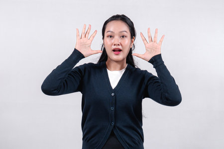 An excited Asian woman in a black cardigan, with long dark hair, holding both hands up with open palms, showing excitement or surprise, against a neutral background.の写真素材