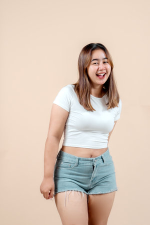 A cheerful young woman smiling widely, standing with her hands behind her back, exuding happiness and confidence against a soft beige background.の写真素材