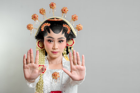 Indonesian bride in Solo Putri attire, wearing an ornate headdress and lace kebaya. She raises both hands in a stop gesture with a serious expression, exuding elegance and grace.の写真素材