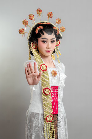 Indonesian bride in Solo Putri attire, wearing a lace kebaya and ornate headdress. She gazes confidently, extending her hand in a stop gesture, exuding elegance and grace.の写真素材