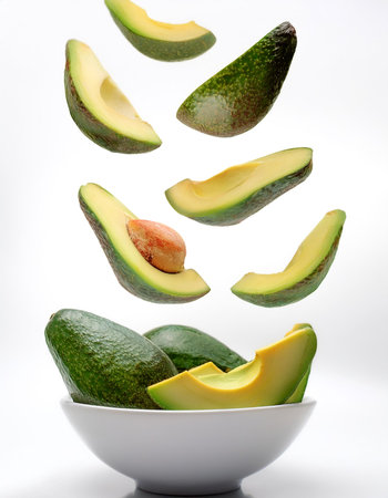 slices or chunks of avocados fruit in a white bowl with chunks or slices floating or flying isolated...