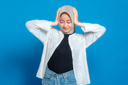 Young Asian Muslim woman in hijab smiling with eyes closed and hands on head. Peaceful facial...
