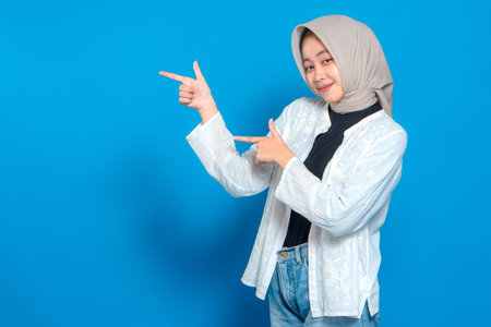 Happy young Asian Muslim woman in hijab smiling and pointing sideways with both hands. Confident facial expression ideal for promotional, advertising, or marketing content.の写真素材