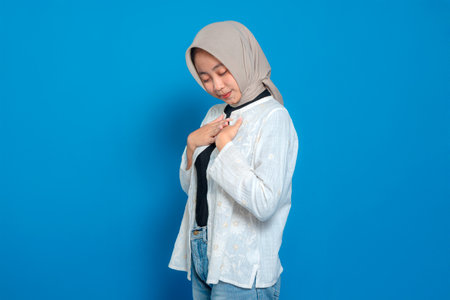 Young Asian Muslim woman in hijab closing her eyes with a peaceful smile, touching her chest gently...