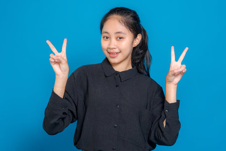 Cheerful young Asian girl smiling brightly while showing double peace signs with both hands. Confident, playful expression perfect for advertising and promotional content.の写真素材
