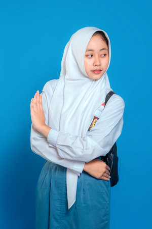 Confident Muslim schoolgirl in uniform and hijab showing stop hand gesture with calm, assertive facial expression. Back to school concept on isolated blue background.の写真素材