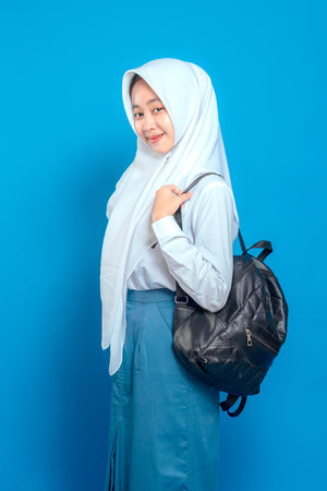 Cheerful Asian teenage girl wearing school uniform and hijab, smiling confidently while holding backpack straps. Positive and friendly facial expression on blue background.の写真素材