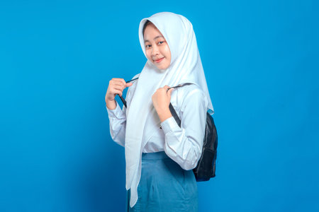 Cheerful Asian teenage girl wearing school uniform and hijab, smiling confidently while holding backpack straps. Positive and friendly facial expression on blue background.の写真素材