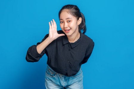 Cheerful young Asian woman smiling and gesturing hello with an open palm near her face, expressing friendliness, happiness, and positivity against a blue background.の写真素材
