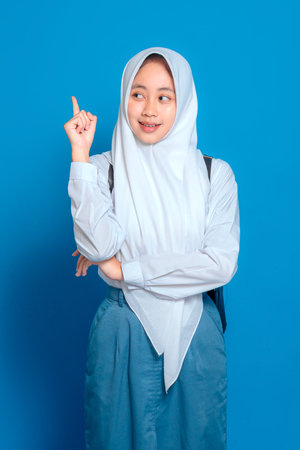 Cheerful Muslim schoolgirl wearing hijab raising finger and smiling, expressing a bright idea or inspiration. Perfect concept for back to school and education themes.の写真素材