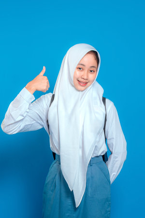 Confident Muslim schoolgirl wearing hijab and uniform giving a thumbs up with a warm smile. Her bright expression conveys optimism, positivity, and encouragement in education.の写真素材