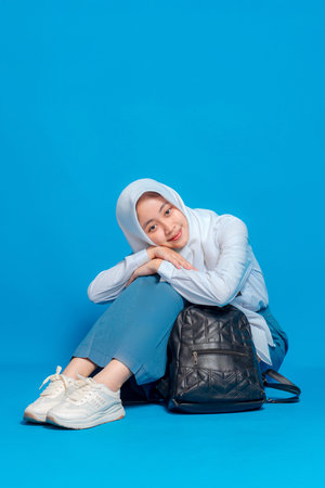 Asian Muslim teenage girl smiling sweetly with a calm and relaxed expression while sitting on the floor and leaning on a black backpack, back to school and education concept.の写真素材