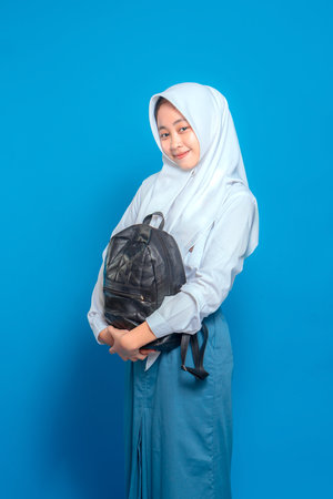 Portrait of a smiling Muslim schoolgirl wearing a white hijab and uniform, holding a black backpack. She looks calm and friendly, standing against a blue background.の写真素材