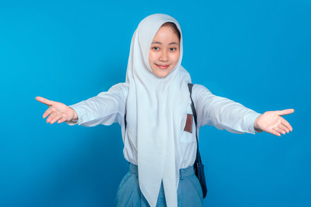 Cheerful Muslim schoolgirl in uniform and hijab smiling with open arms, welcoming warmly. Her happy facial expression conveys friendliness. Isolated on blue background..の写真素材